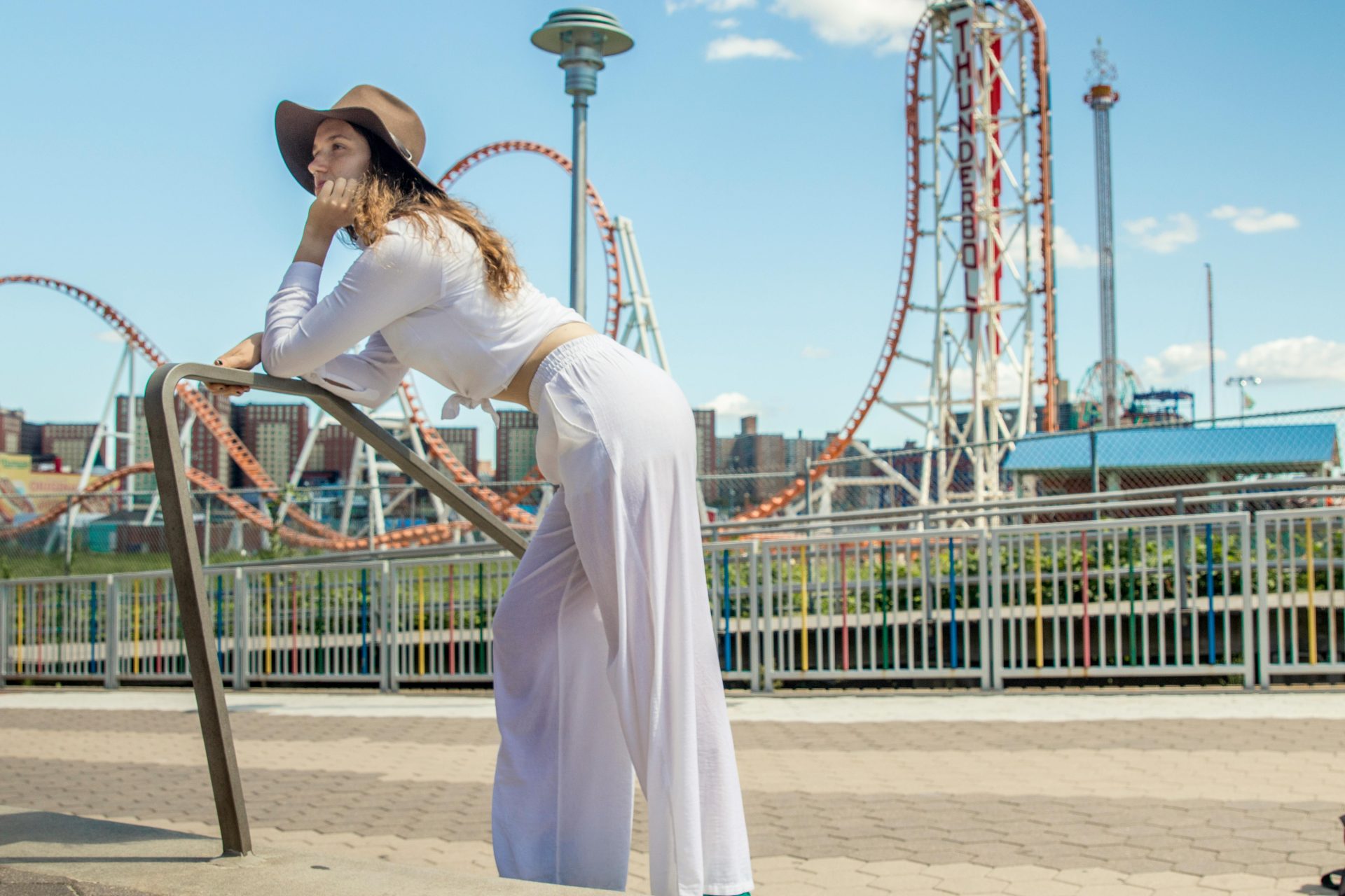 Modische Frau posiert an einem sonnigen Tag in einem Vergnügungspark neben einer Achterbahn.